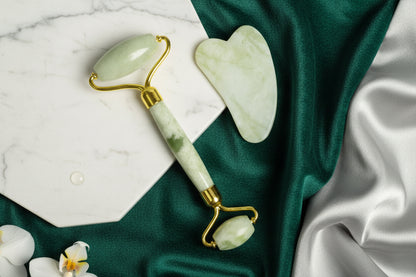 Jade roller and gua sha tool on a marble surface with green fabric and flowers.